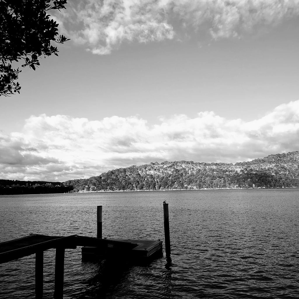 Black and white landscape view of a jetty on calm waters framed by forested hills, reflecting the natural Australian coastal inspiration behind B108 Design.