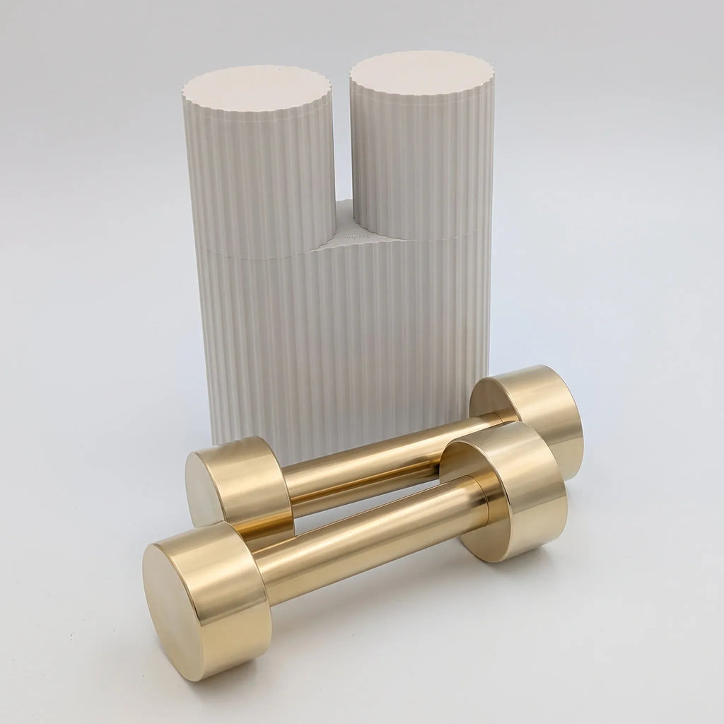 Studio shot of a pair of solid brass hand weights lying in front of their custom off-white ribbed pillar packaging by B108 Design.